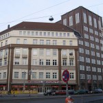 Mehrstöckiges Eckhaus mit beiger Fassade und braunen Akzenten, davor ein rotes Verkehrsschild – Jobvermittlung bei Staff Eins.