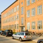 Backsteingebäude mit großen Fenstern, davor geparkte Autos und Fahrräder – Jobvermittlung bei Staff Eins.